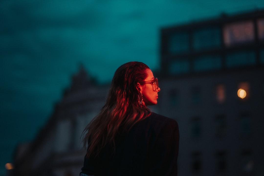 A person with long hair and glasses stands under a vibrant, moody sky, surrounded by buildings. The image captures an urban night scene.